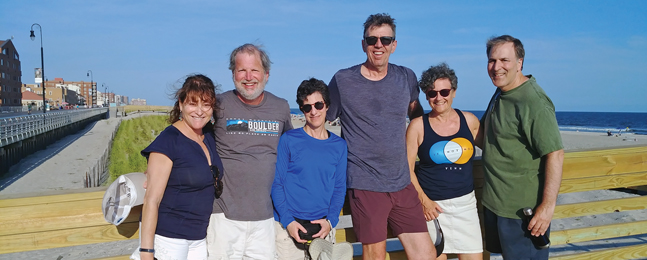 Three men and three women dressed in summer clothes pose with a beach and a boardwalk in the background.